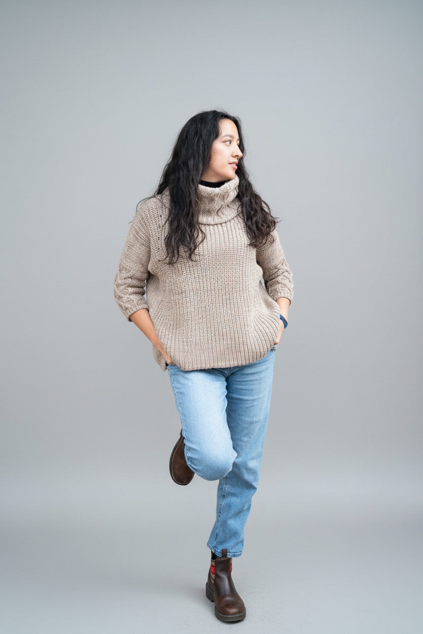 Woman wearing a beige sweater and blue jeans leaning against a brick wall.