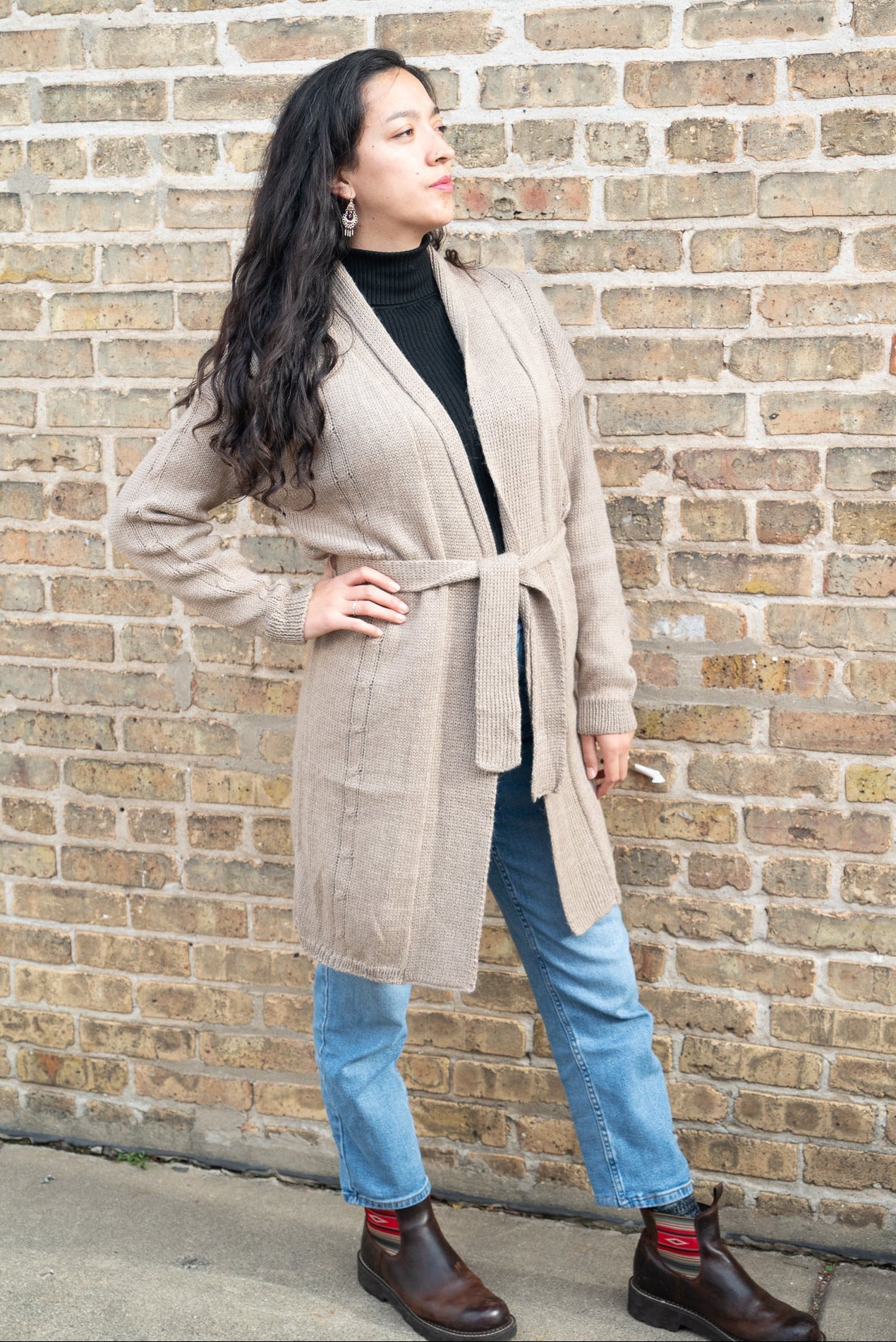 Woman wearing a beige cardigan, black top, blue jeans, and brown shoes against a brick wall.